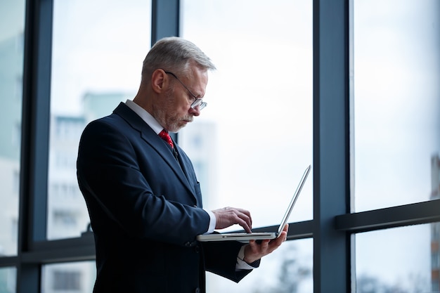 Smiling happy managing director thinks about his successful career development while standing with a laptop in his office near the background of a window with copy space