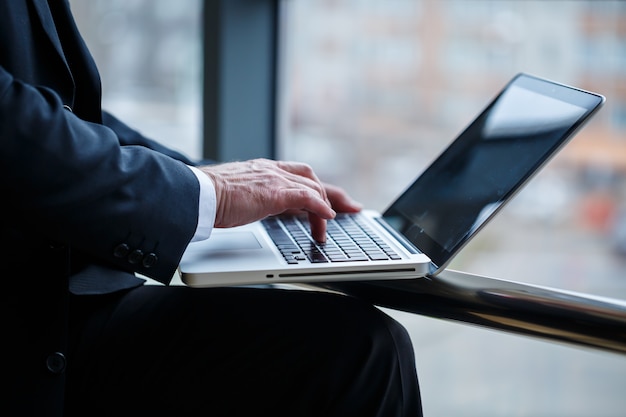 Smiling happy managing director thinks about his successful career development while standing with a laptop in his office near the background of a window with copy space