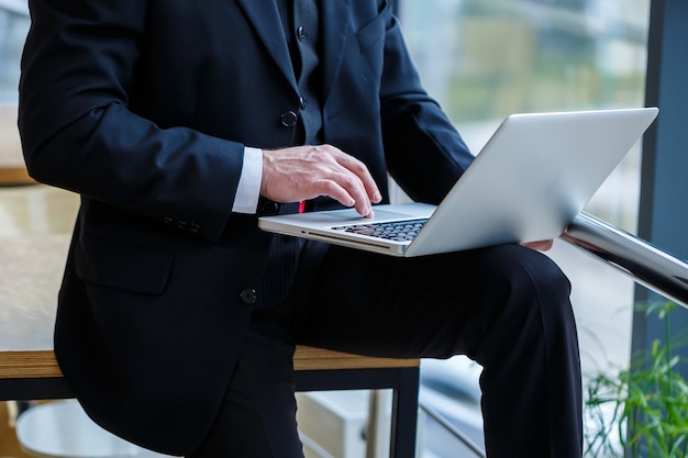 Smiling happy managing director thinks about his successful career development while standing with a laptop in his office near the background of a window with copy space