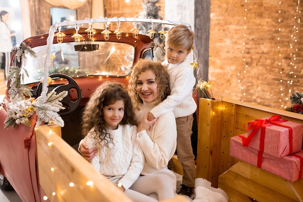 Smiling happy mother with her son and daughter in red car pickup trailer near Christmas trees and lights