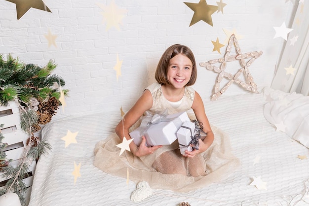 Smiling happy teenage girl in a smart dress with New Year's gifts sits on a bed in a Christmas interior Traditional family holidays