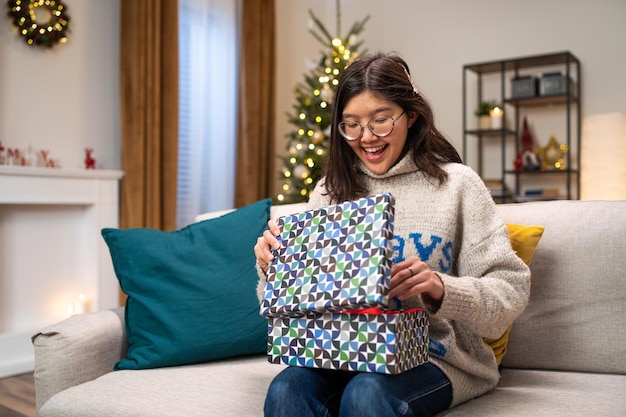 A smiling happy woman in glasses and a winter sweater sits on a couch in a cozy living room