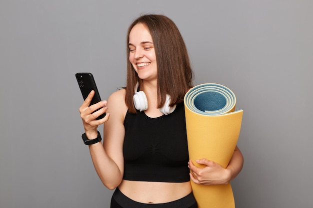 Smiling happy woman holding rolled fitness mat dressed in activewear isolated over gray background using modern mobile phone downloads application for sport training