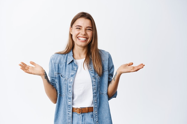 Smiling happy woman spread hands sideways and laughing, have nothing to say, standing against white wall