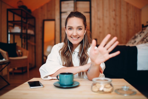 Smiling happy young woman sitting at table at home behind computer laptop and talking on video call Girl female with cup of tea or coffee speaking online on webcam indoors