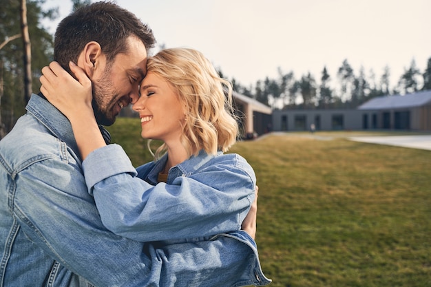 Smiling high-spirited attractive blonde female and her boyfriend in denim jackets hugging each other outside