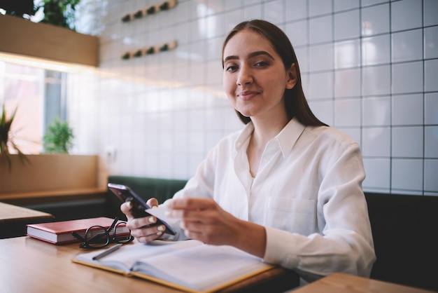 smiling hipster girl sitting at coffee shop looking at camera and holding business card in hand