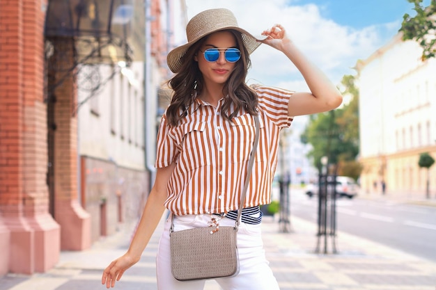 Smiling hipster trendy girl posing at the city summer streetholding hat