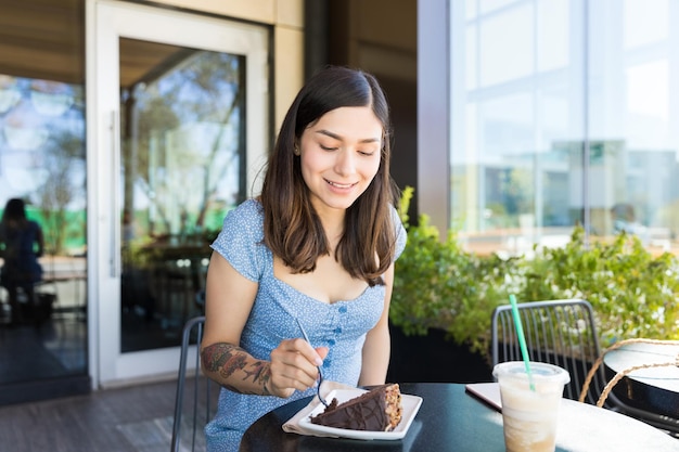 Smiling Hispanic brunette eating slice of cake at while sitting at cafe