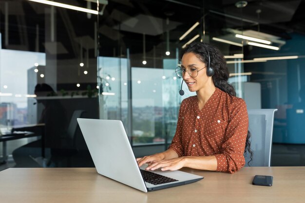 Smiling hispanic businesswoman working inside office with laptop and headset for video call woman