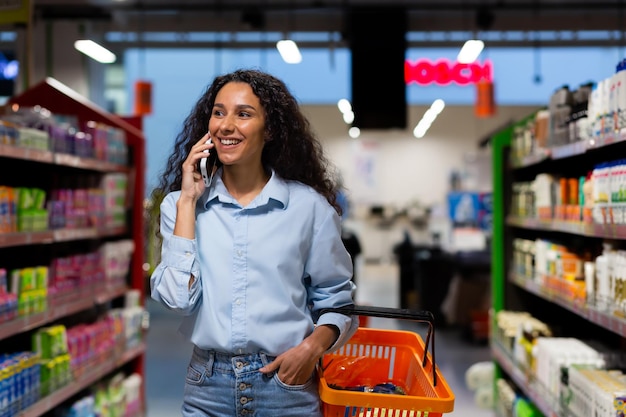 Smiling hispanic woman shopper in supermarket walks between rows of shelves with goods woman