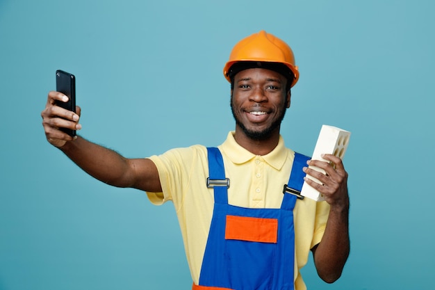 Smiling holding brick young african american builder in uniform take a selfie isolated on blue background