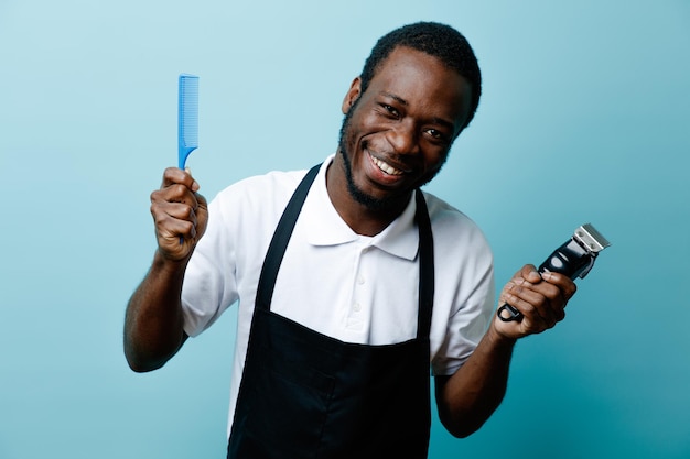 Smiling holding comb with hair clipper young african american barber in uniform isolated on blue background