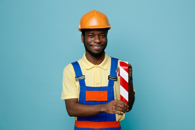 Smiling holding duct tape young african american builder in uniform isolated on blue background