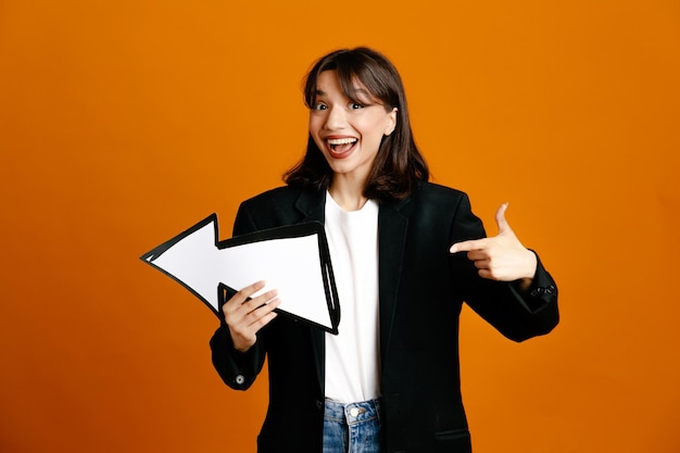 smiling holding and points at direction mark young beautiful female wearing black jacket isolated on orange background