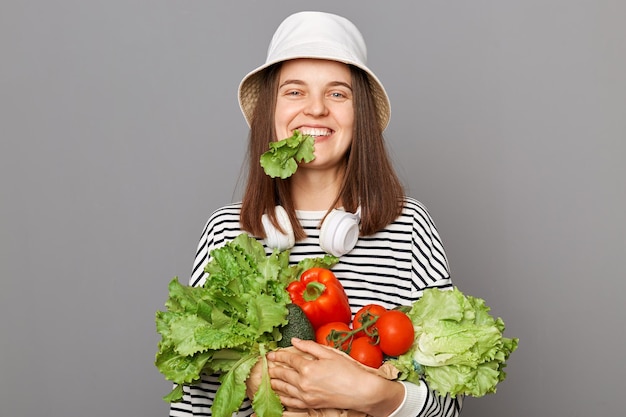 Photo smiling hungry caucasian woman wearing striped shirt and panama holding organic vegetables standing isolated over gray background keeps diet eating green lettuce