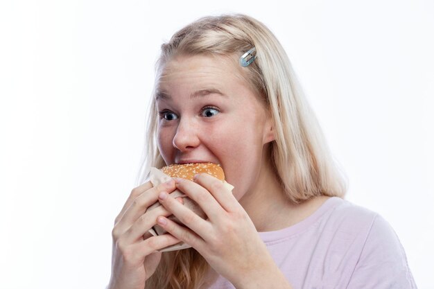 Smiling hungry teenage girl takes a bite of hamburger Delicious and popular fast food A cute blonde with freckles in a pink jacket White background