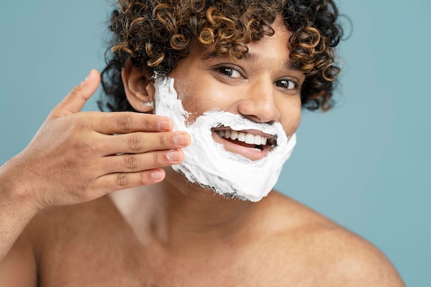 Smiling Indian man applying shaving foam on his face, looking at mirror in bathroom. Skin care