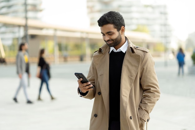 Smiling Indian man holding mobile phone reading text message, communication standing on urban street