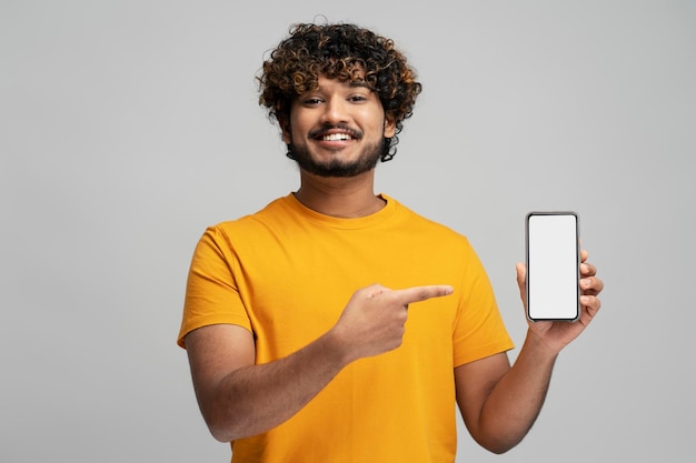 Smiling Indian man holding mobile phone, shopping online pointing finger at empty display. Mockup