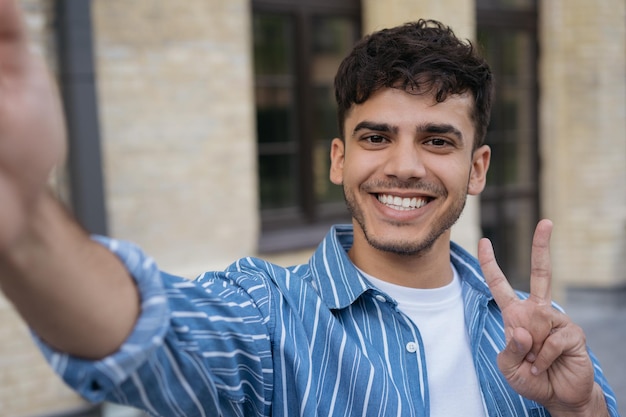 Smiling Indian man holding mobile phone taking selfie showing victory sign on the street