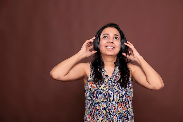 Smiling indian woman listening to music in wireless headphones with satisfied facial expression. Lady wearing earphones, enjoying song, looking upwards, front view studio medium shot