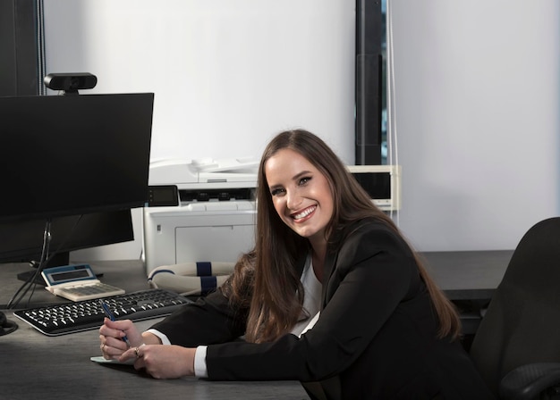 Smiling Israeli businesswoman sitting on table in the office and write on book Caucasian woman with long hair dressed black suit in office working on desktop computer writing notes in her day