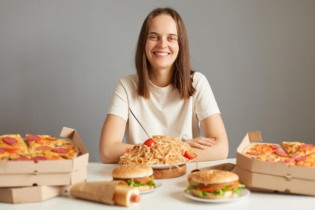 Smiling joyful young caucasian woman with brown hair wearing white Tshirt sitting at table among fast food isolated over gray background likes to eat unhealthy dishes