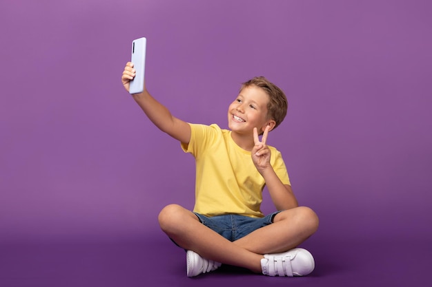 Smiling kid boy preteen looking to the cell phone taking selfie Cheerful child using mobile phone sitting on the floor on purple background Studio shot