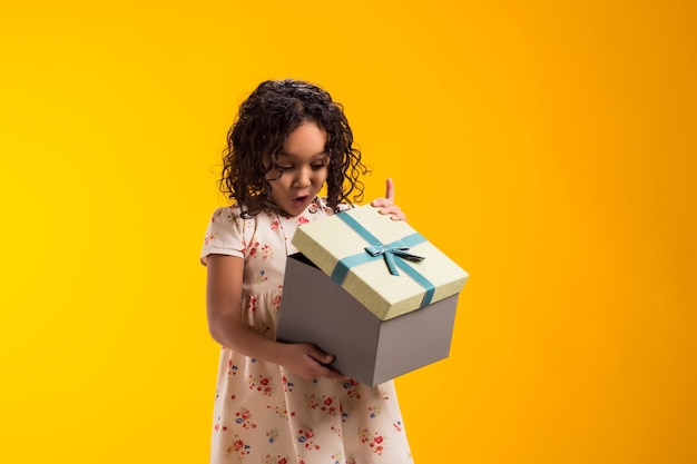 Smiling kid girl holding giftbox and looking inside the box over yellow background Birthday and celebration concept