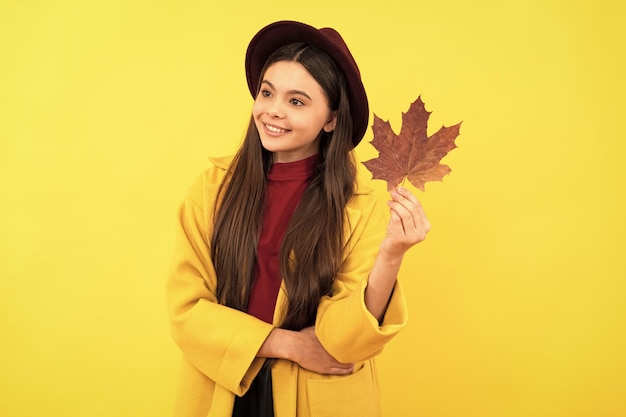 Smiling kid in hat and coat with autumn maple leaf on yellow background season