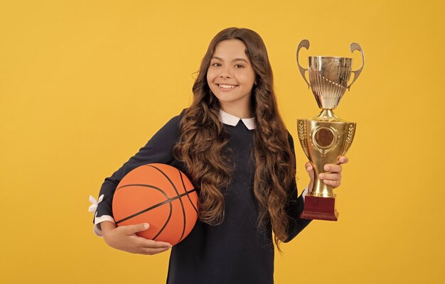 Smiling kid hold basketball ball and champion cup on yellow background achievement