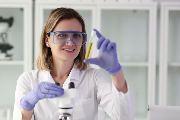 Smiling laboratory assistant in protective glasses examines sample of yellow liquid in test