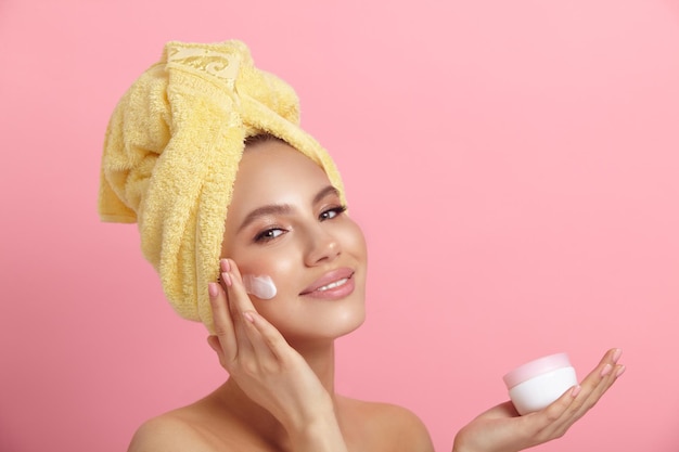 Smiling lady applies cream on face showing jar on pink