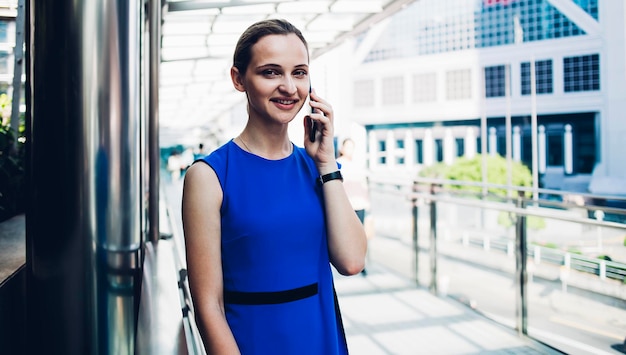 Smiling lady in blue stylish sleeveless dress making call standing on covered terrace in sunny day