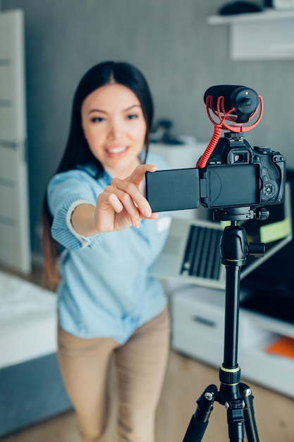 Smiling lady with a laptop in hand leaning towards the camera on a tripod while adjusting it