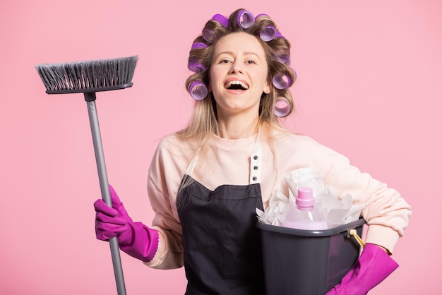 A smiling laughing girl with rollers in hair looks into the camera contentedly holding a broom and mop bucket Woman gets ready to mop the floor of the apartment pink studio background