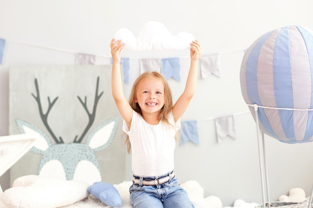 Smiling little blonde girl holding a cloud pillow  of a decorative balloon. The child plays in the children's room with toys. The concept of childhood, travel. Toddler in kindergarten