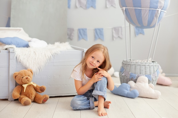 Smiling Little blonde girl plays in a children's room with teddy bear. Child in kindergarten plays in  with a toy.