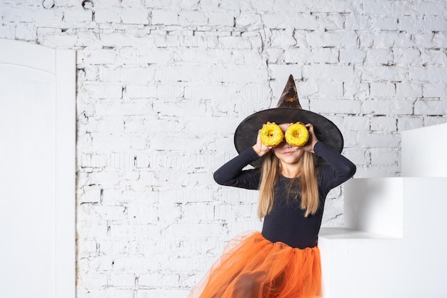 A smiling little blonde girl in a witch costume holding yellow donuts in her hands sitting on the steps of the house