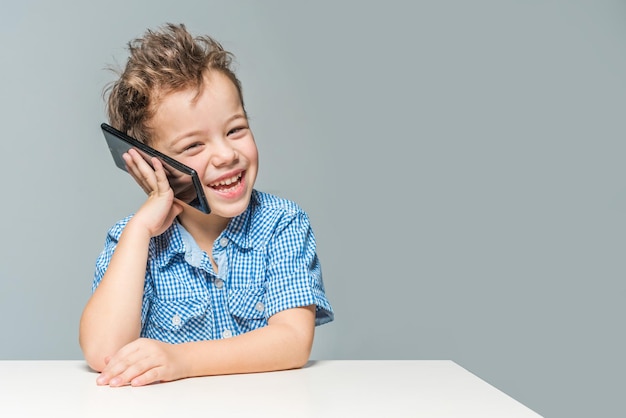 Smiling little boy talking on the phone while sitting at the table on a pink background Toned