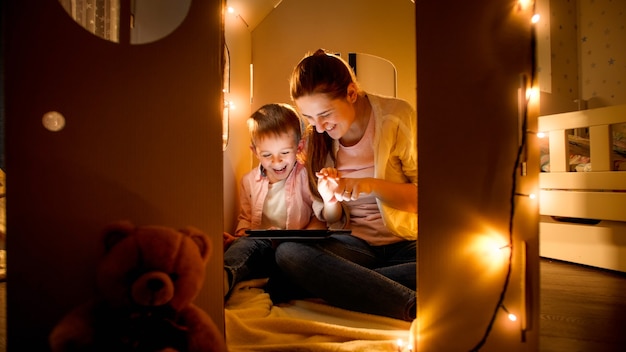 Smiling little boy with mother playing in toy cardboard house and using tablet computer. Concept of child education and family having time together at night.