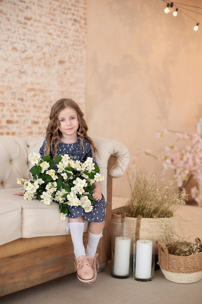 Smiling little curly-haired girl with a bouquet of jasmine flowers sits on the sofa in a cozy living room.