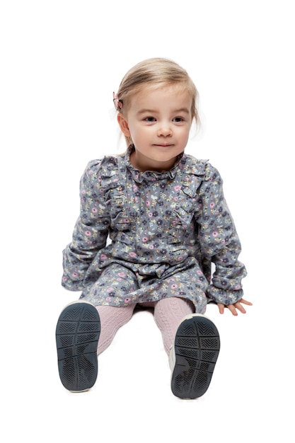 Smiling little girl 3 years old is sitting on the floor. Simpachy child in a flowered dress. Love and tenderness. Isolated on white background. Vertical.