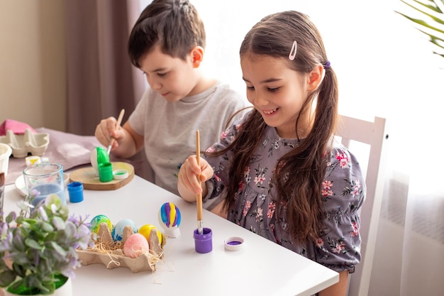 A smiling little girl and boy sits at a white table near the window paints eggs