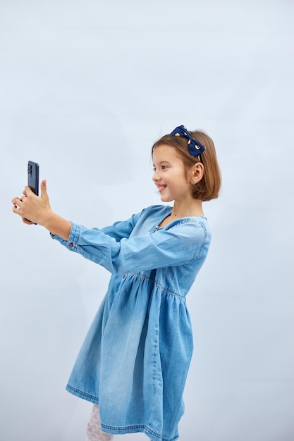 Smiling little girl in casual denim dress hold smartphone do selfie