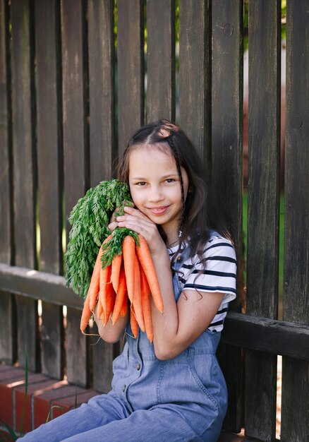 Smiling little girl in a garden holds a bunch of fresh carrots Vertical