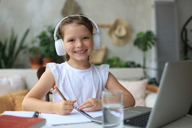 Smiling little girl in headphones handwrite study online using laptop at home, cute happy small child in earphones take Internet web lesson or class on PC