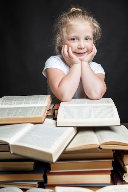 Smiling little girl is sitting with a pile of books Education and training of children Black background Vertical