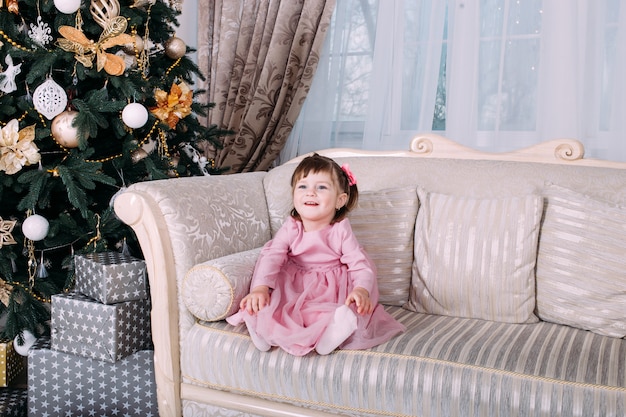 Smiling little girl sitting on sofa near Christmas tree indoors. Merry Christmas and Happy New Year!